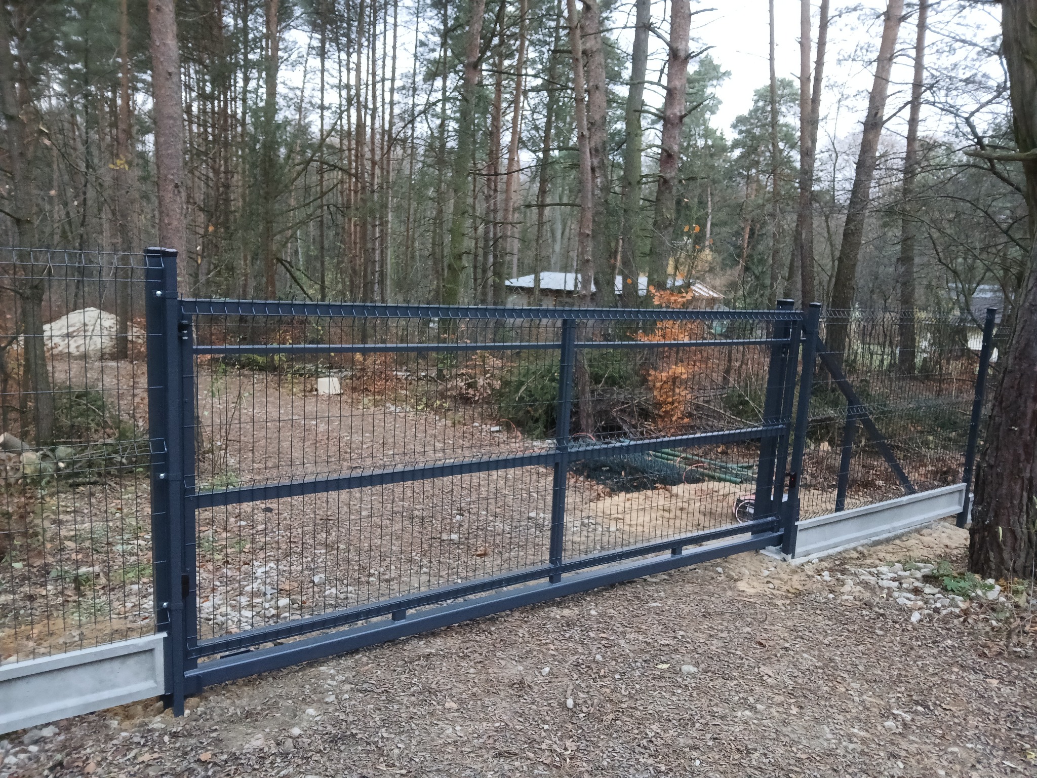 Ciemnoszara brama przesuwna z paneli ogrodzeniowych, zamontowana na betonowej podmurówce, na tle leśnego krajobrazu z widocznym domem w oddali.