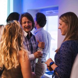 zofia.jpg Zofia Kowalczyk - Uśmiechnięty mężczyzna w koszuli w kratę trzyma butelkę podczas rozmowy z kobietą na imprezie, otoczony innymi osobami w pomieszczeniu z niebieskim oświetleniem.