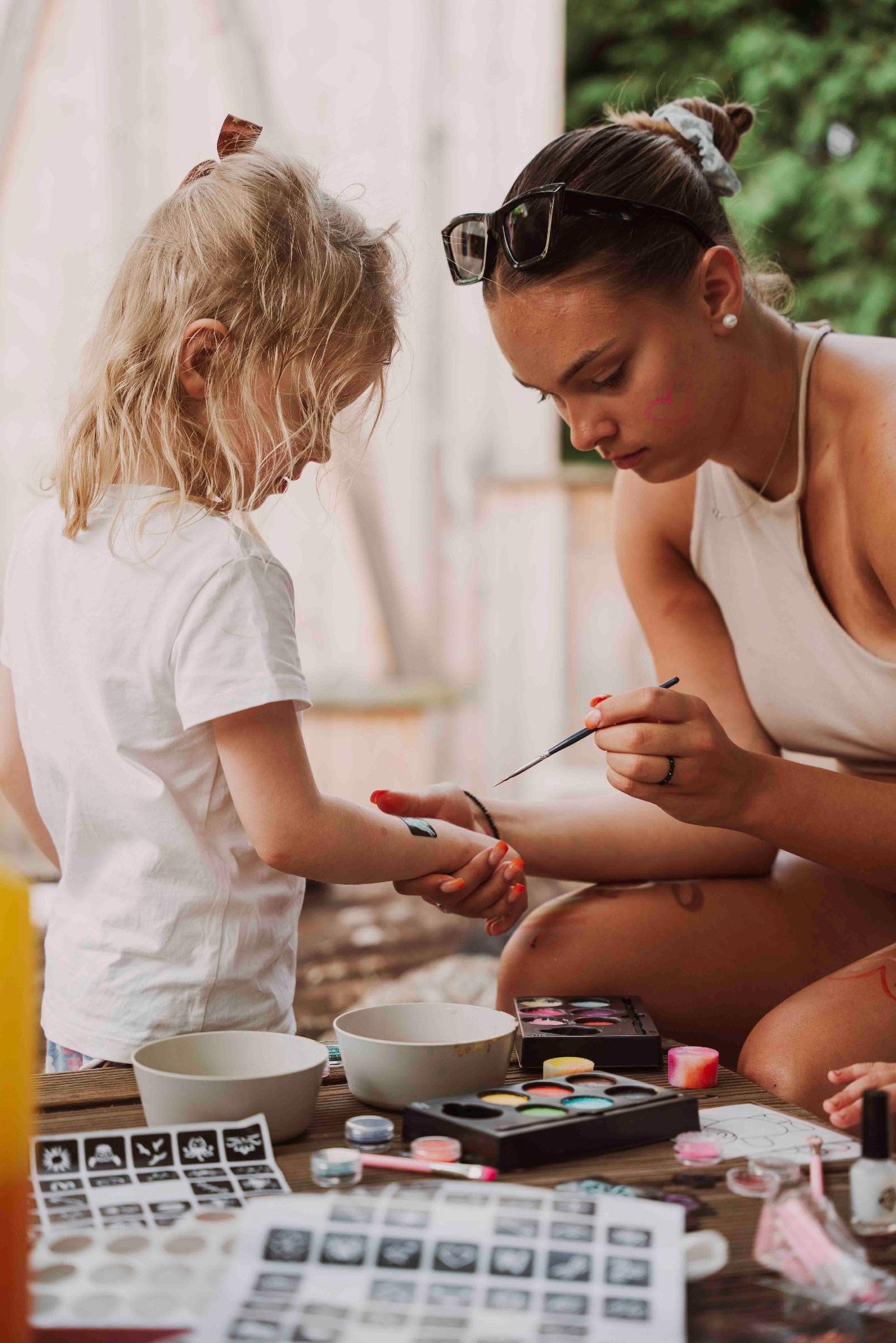 Kobieta maluje pędzelkiem wzór na ramieniu dziecka. Na stole farby, stencils, miseczki. Ujęcie z bliska, pokazujące detale malowania.