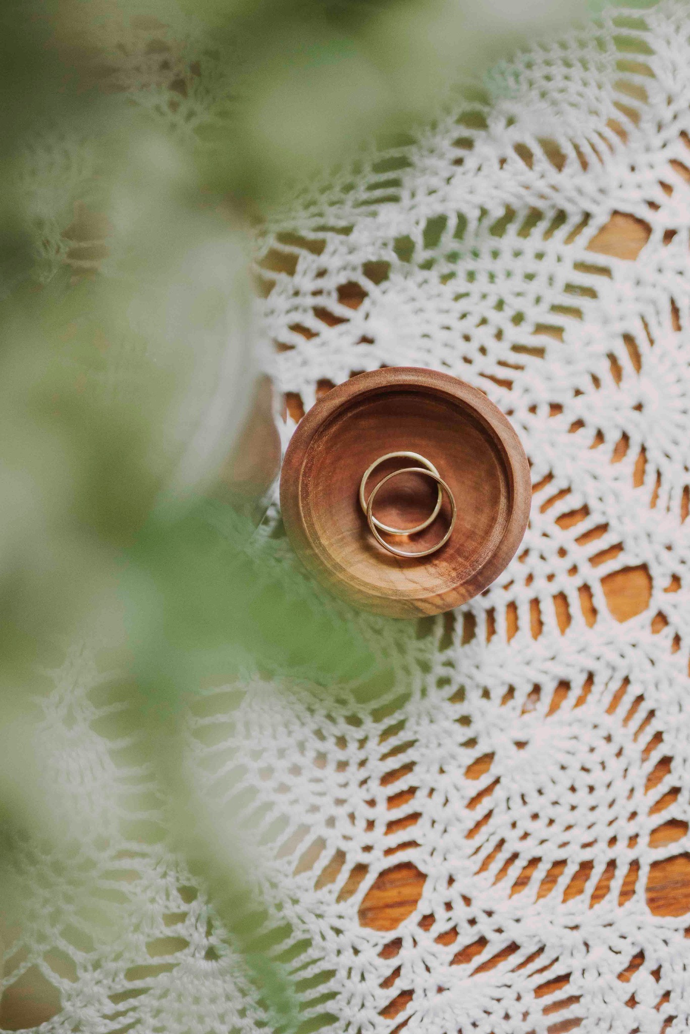 Dwa złote obrączki ślubne w drewnianej miseczce na białej, koronkowej serwetce, z rozmytym zielonym tłem roślinnym po lewej stronie. Delikatne, naturalne światło.