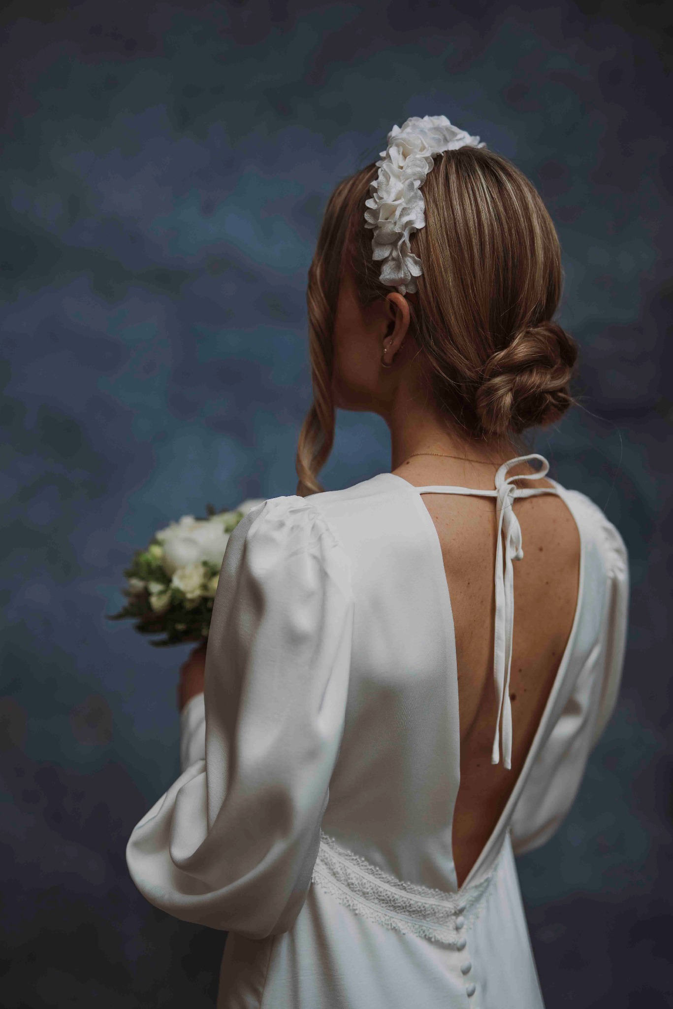 Panna młoda z tyłu, w białej sukni z odkrytymi plecami i kwiatowym wiankiem, trzyma bukiet na rozmytym, ciemnym tle. Delikatne oświetlenie podkreśla detale sukni.