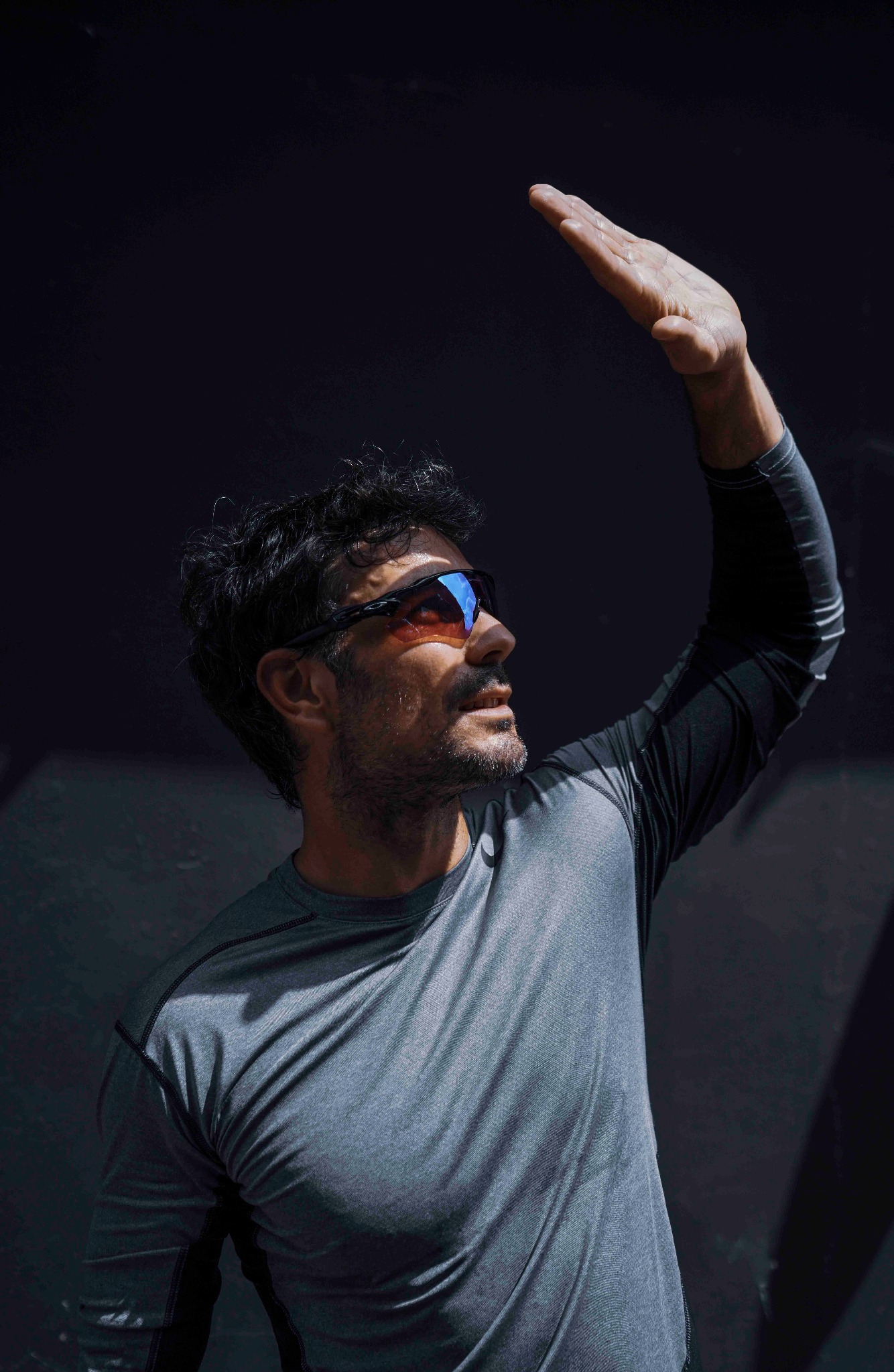 Mężczyzna w okularach przeciwsłonecznych, w szarej koszulce z długim rękawem, zasłaniający się dłonią przed słońcem na ciemnym tle. Ujęcie od dołu.