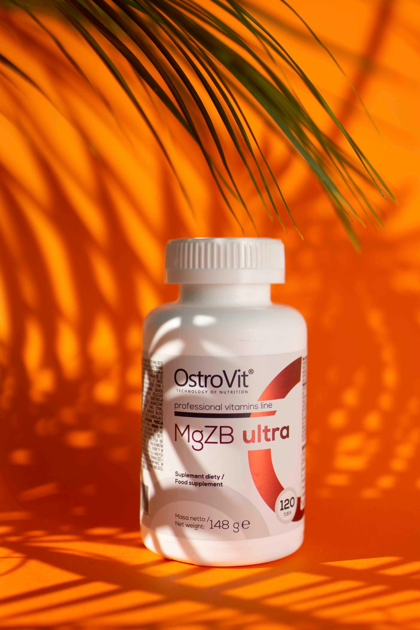 Białe opakowanie suplementu diety OstroVit MgZB ultra, 120 tabletek, na pomarańczowym tle z zielonym liściem palmy w tle. Widoczne informacje o wadze netto: 148 g.