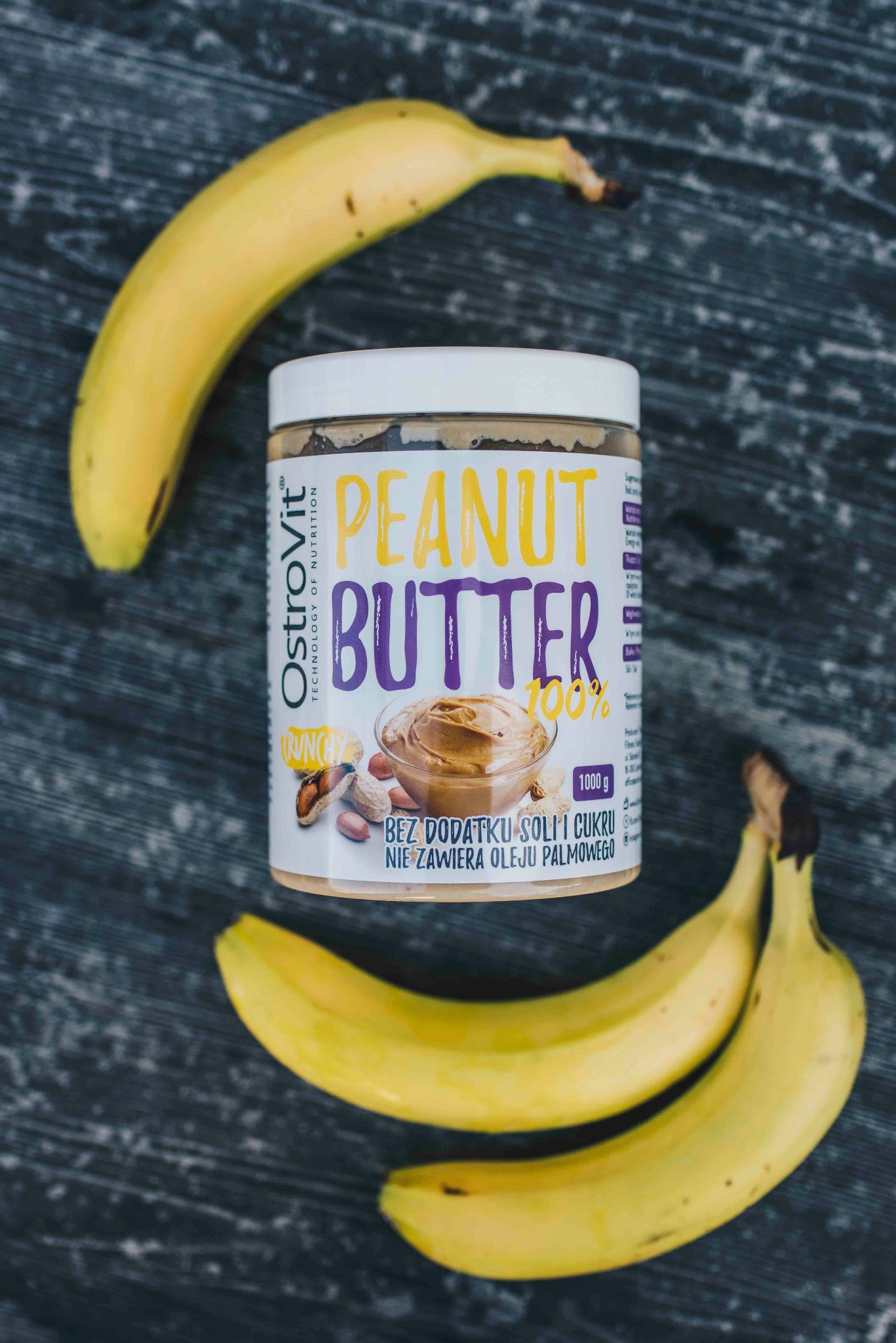 Słoik masła orzechowego Ostrovit 100% crunchy z bananami na ciemnym, teksturowanym tle. Fotografia produktowa w Warszawie, z bliska, dobre oświetlenie.
