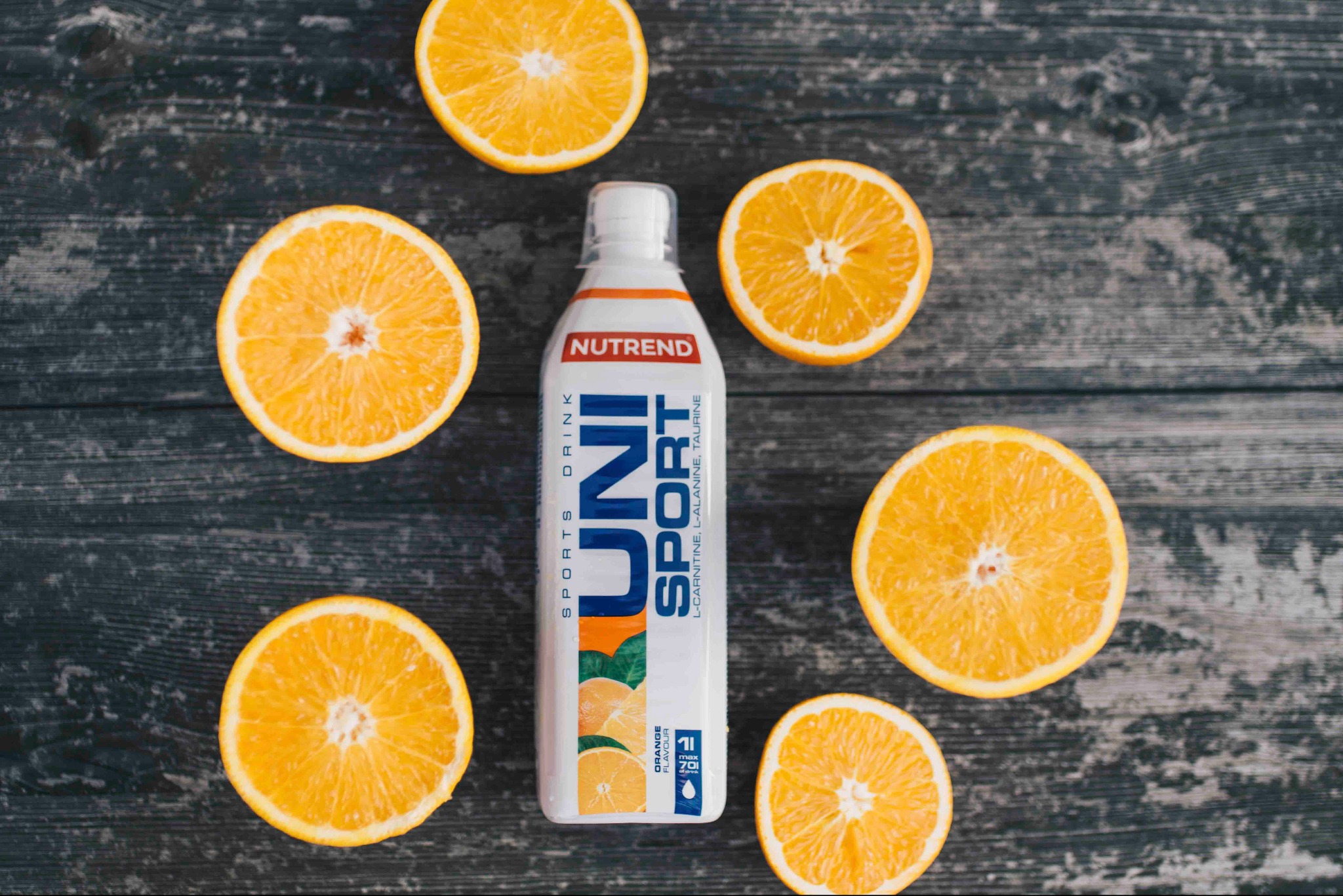Biała butelka napoju izotonicznego Nutrend Uni Sport otoczona połówkami pomarańczy na ciemnym, drewnianym tle. Widok z góry, ostre światło.