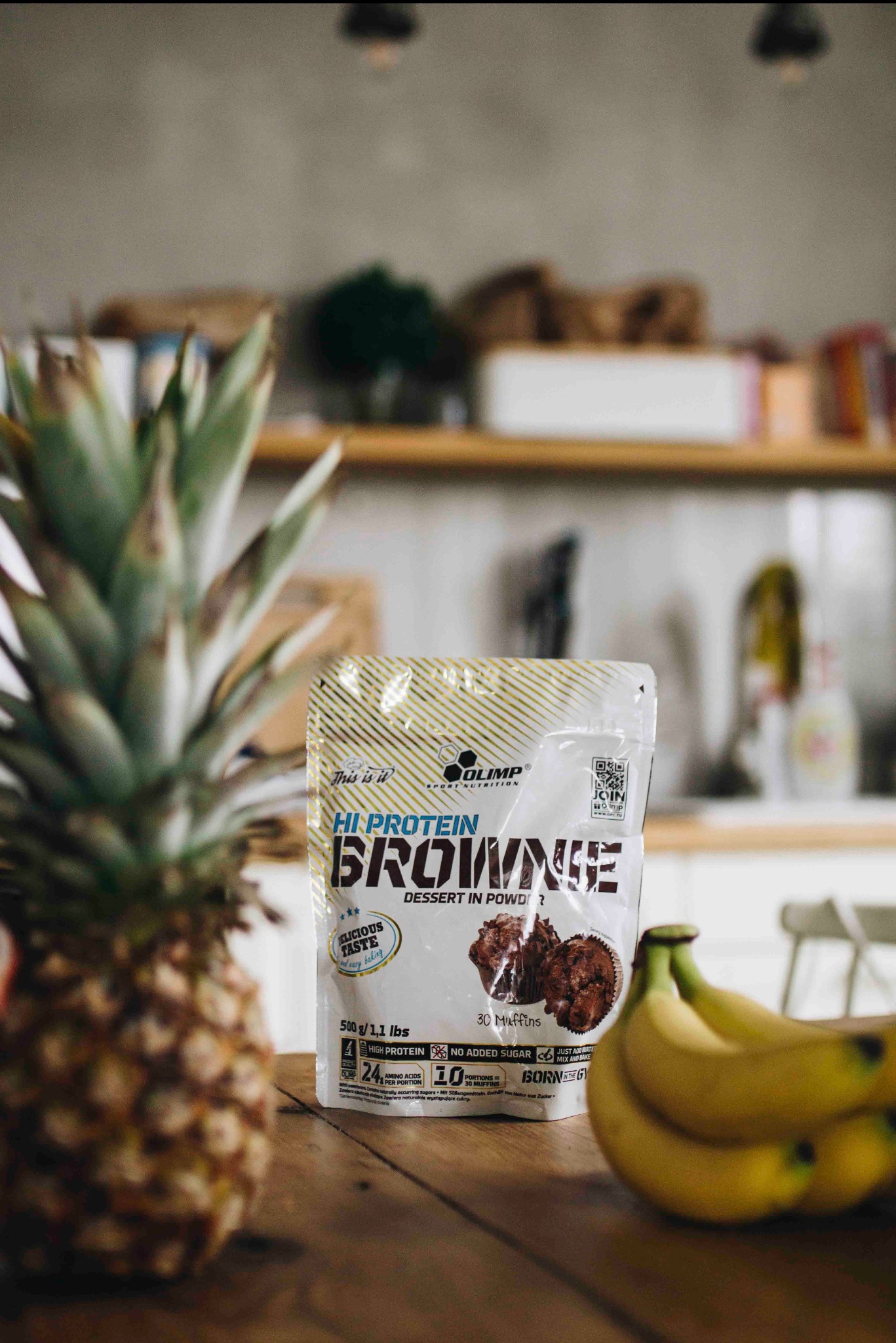 Hi-Protein Brownie w proszku marki Olimp, ananas i banany na drewnianym stole w kuchni. Zdjęcie produktowe z bliska, oświetlone naturalnym światłem.