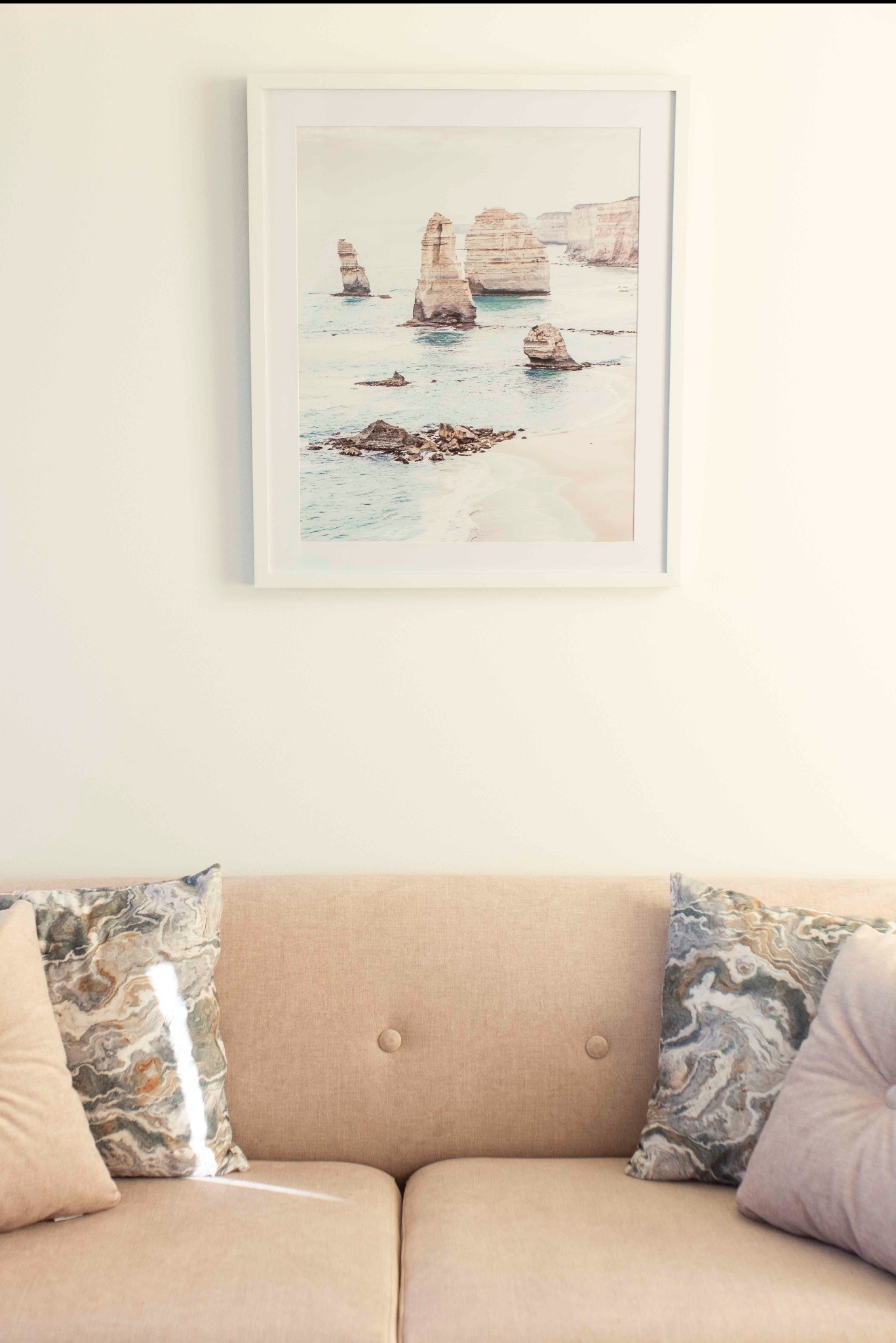 Jasny salon: beżowa sofa z poduszkami pod obrazem w białej ramie, przedstawiającym formacje skalne w oceanie. Minimalistyczny wystrój, stonowana kolorystyka.