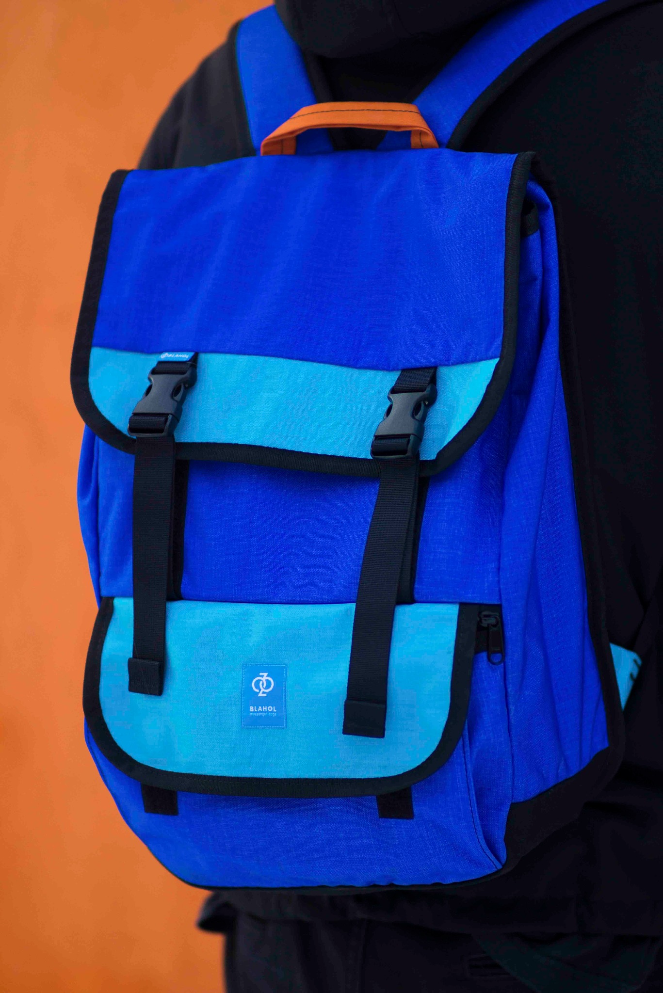 Niebieski plecak miejski z czarnymi klamrami i pomarańczowym uchwytem, widoczny na sylwetce osoby na tle pomarańczowej ściany. Detal produktu z logo marki.