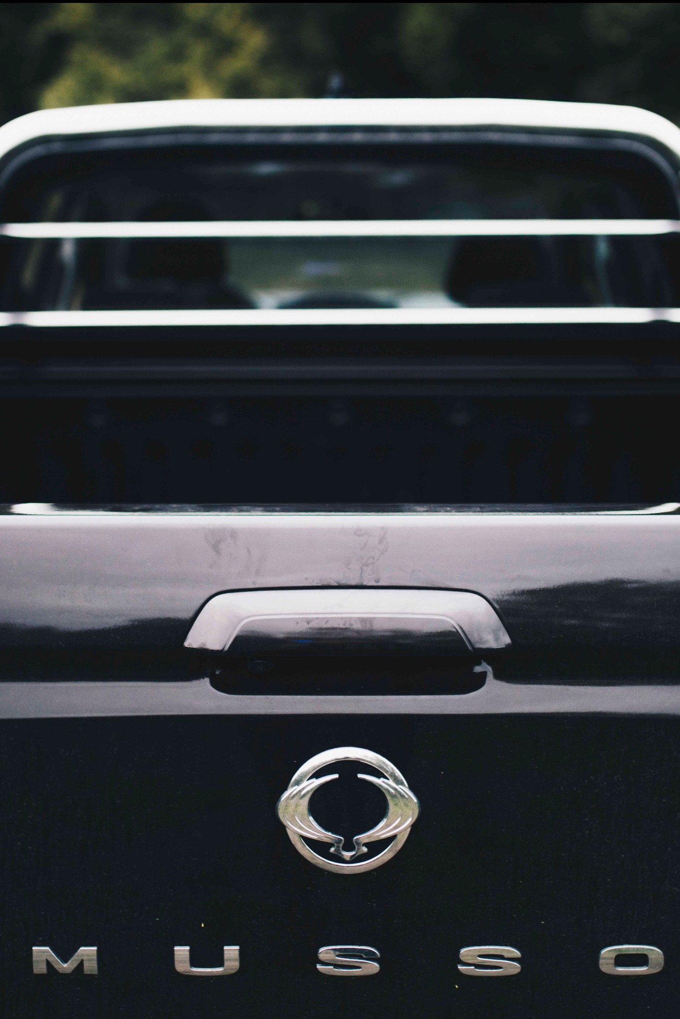 Ciemny samochód marki Musso z chromowanym logo i napisem, widok z tyłu, oświetlenie naturalne, lekki cień. Detal bagażnika i tylnych świateł.