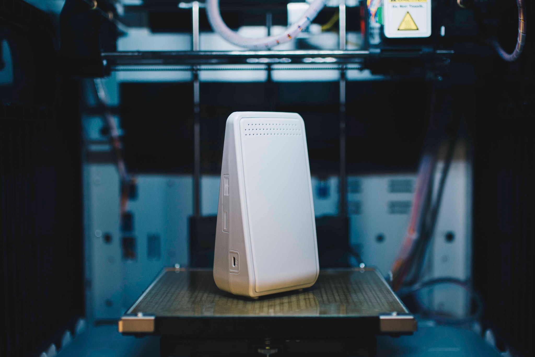 Biały, trójkątny model urządzenia z otworami, wydrukowany na drukarce 3D. Widoczne detale drukarki, kabel zasilający i ostrzeżenie.