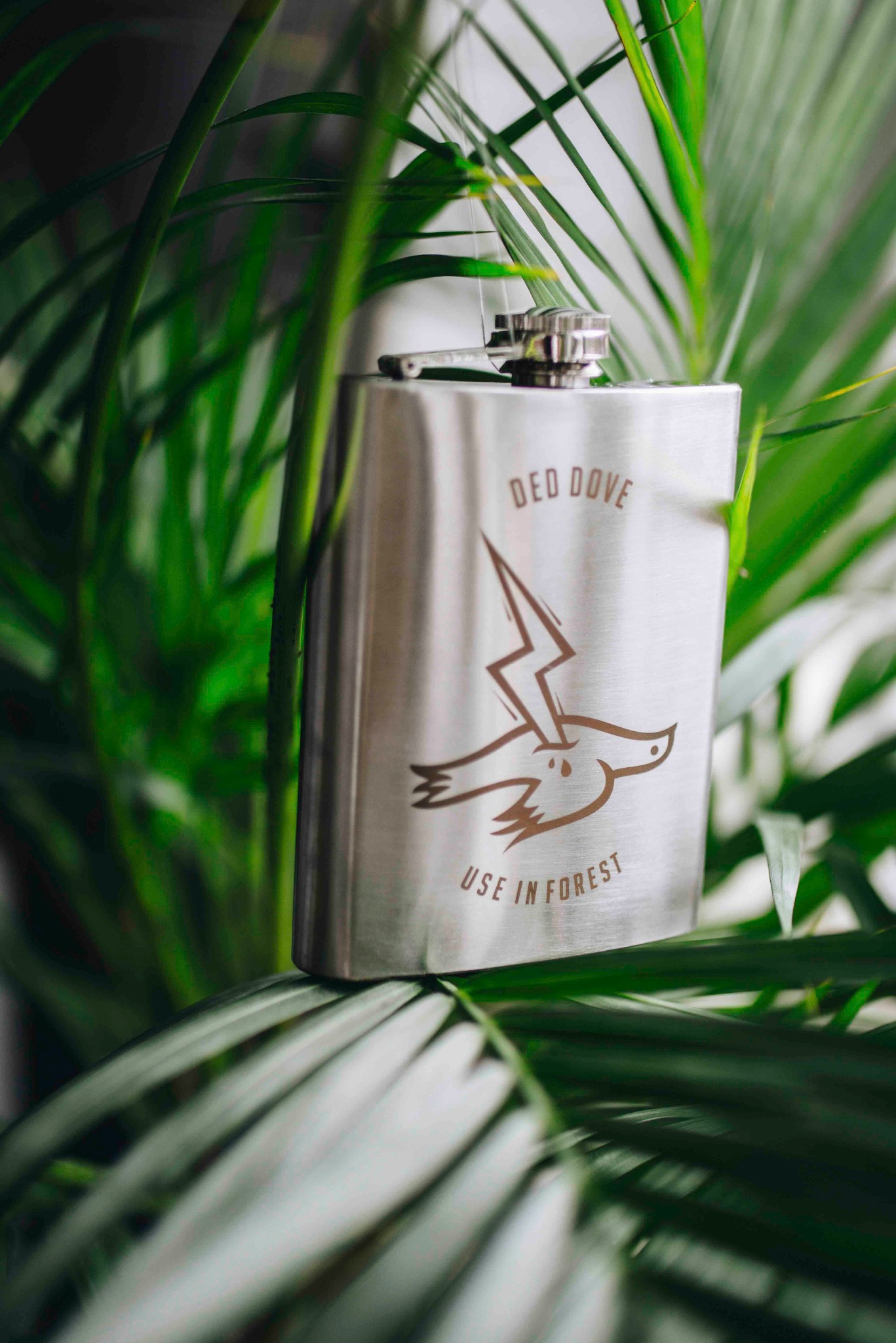 Metalowa piersiówka z logo 'Red Dove' i napisem 'Use in Forest' na tle zielonych liści palmy. Minimalistyczny design, naturalne światło, bliskie ujęcie.