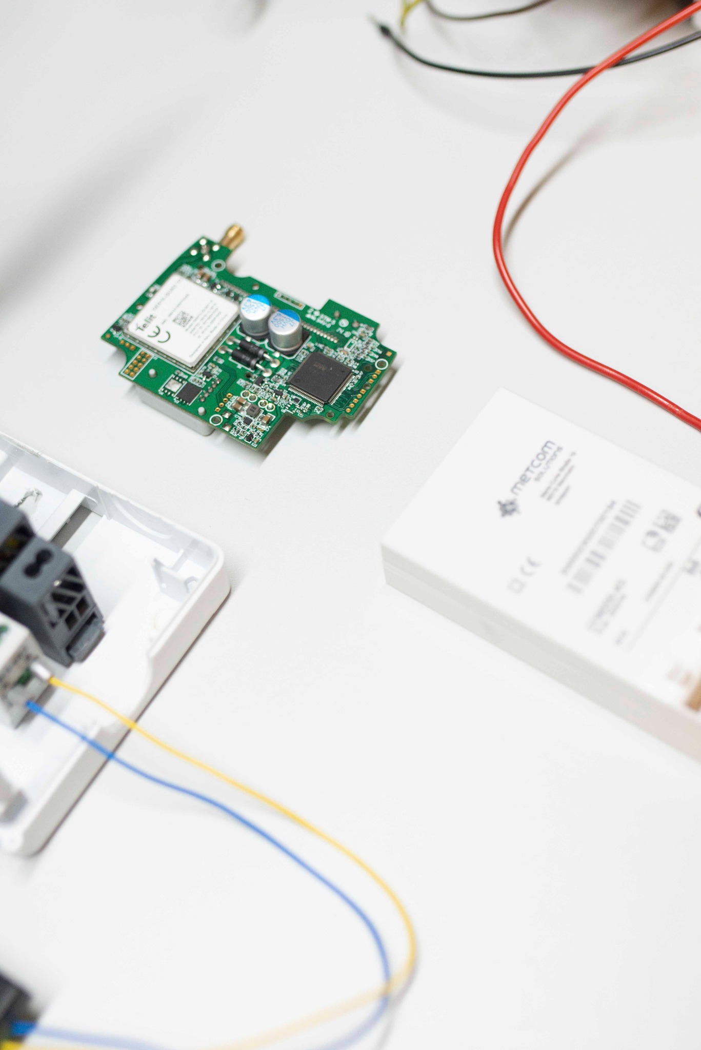 Płytka drukowana z elektroniką Telit, obudowa Metcom, przewody wpięte w złączki na białym tle. Widok z góry, bliski plan.