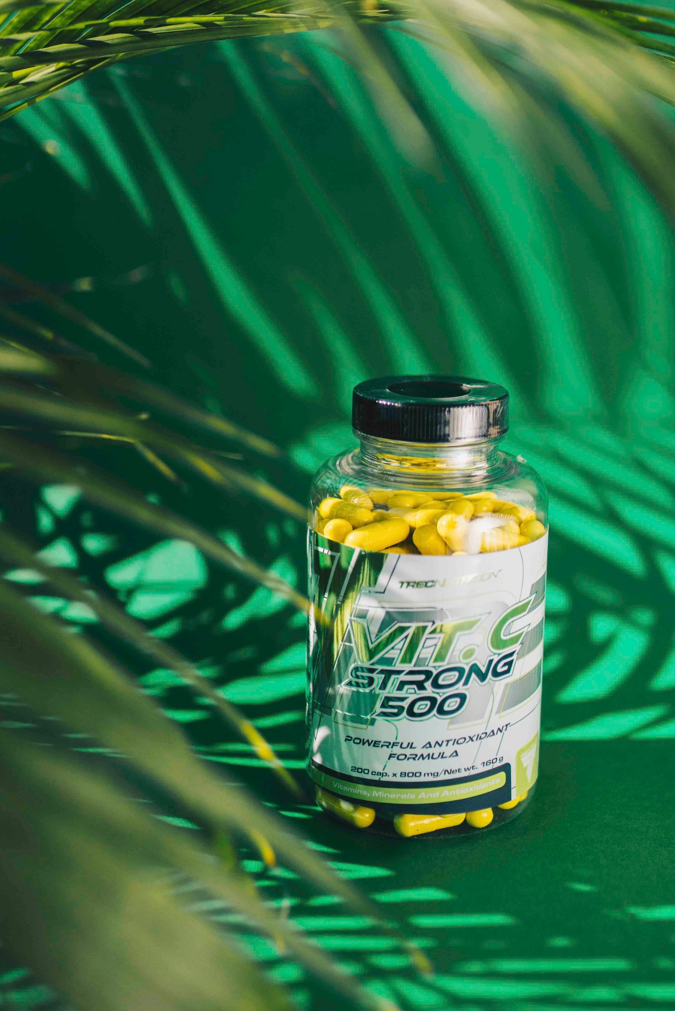 Butelka witaminy C Strong 500 na zielonym tle z cieniem liści palmowych. Produkt Trecnutrition w przezroczystym opakowaniu.