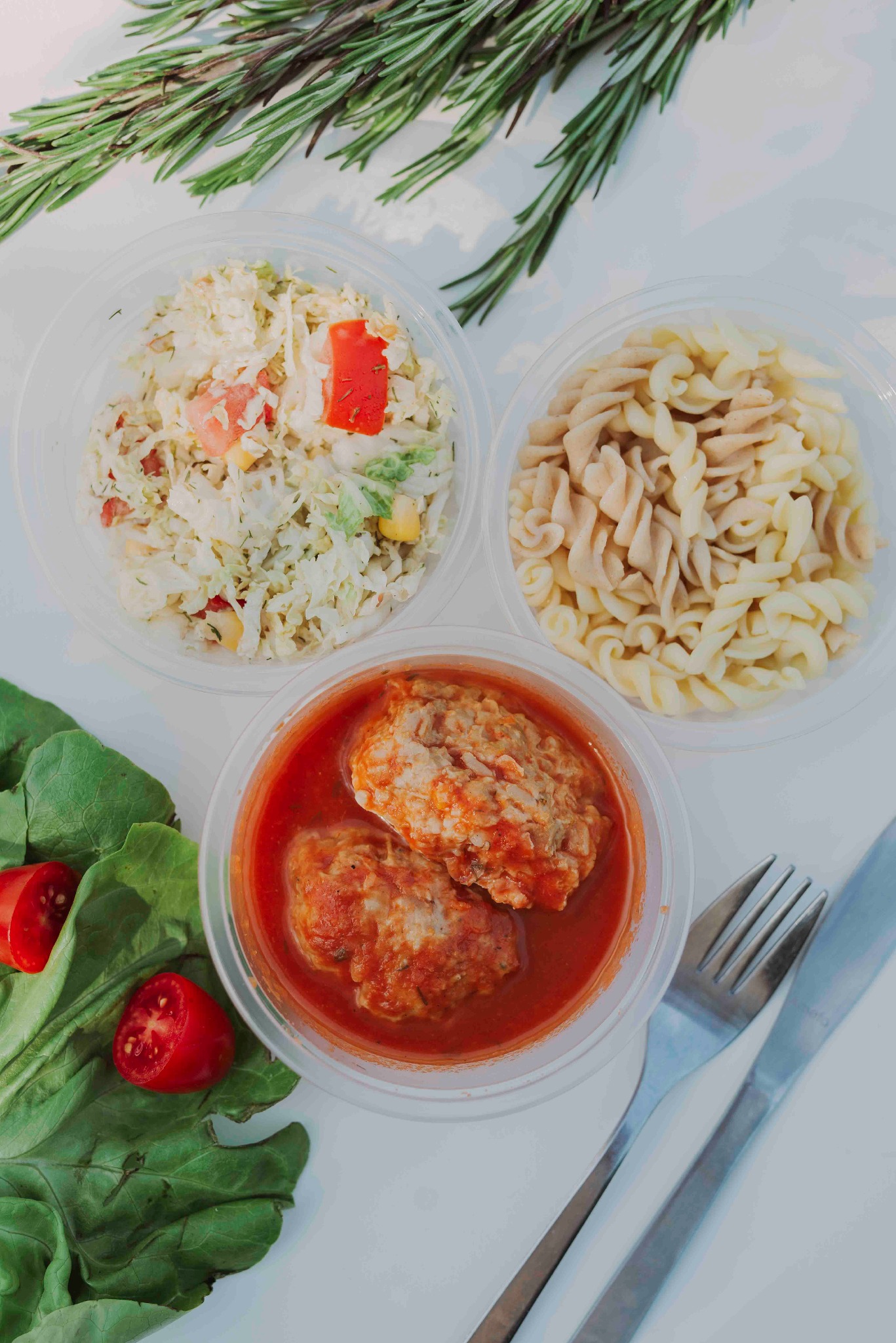 Zestaw obiadowy w plastikowych pojemnikach: sałatka z kapusty, makaron świderki i klopsiki w sosie pomidorowym, uzupełnione świeżym rozmarynem i liśćmi sałaty.