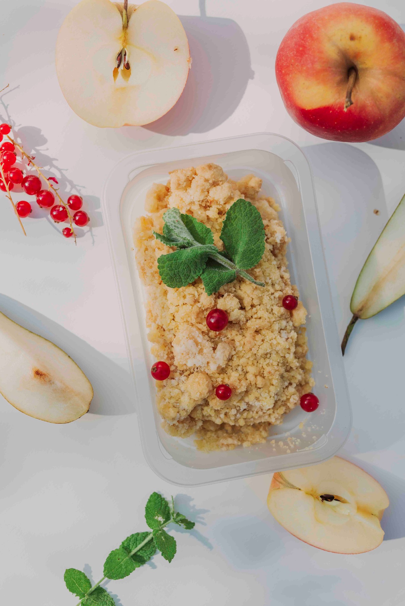 Kruche ciasto w pojemniku z jabłkami, gruszkami i czerwoną porzeczką na białym tle, udekorowane miętą. Naturalne światło, widok z góry.