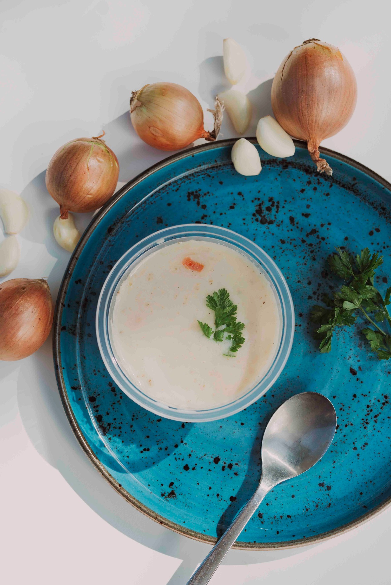 Kremowa zupa w plastikowej miseczce na turkusowym talerzu, otoczona cebulą, czosnkiem i natką pietruszki. Srebrna łyżka obok. Ujęcie z góry w jasnym świetle.