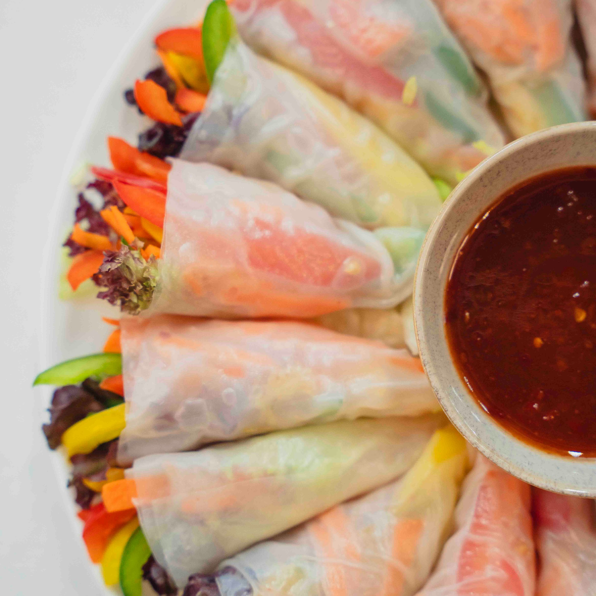 Aromatyczne sajgonki z warzywami i sosem w miseczce, ułożone na talerzu. Widoczne: marchew, papryka, sałata. Jasne tło, naturalne światło, zbliżenie na detale.