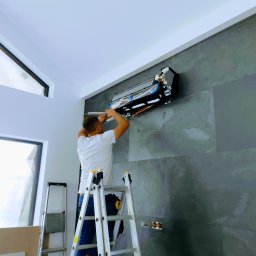 GOWENT SPÓŁKA Z OGRANICZONĄ ODPOWIEDZIALNOŚCIĄ - Instalator na drabinie podłącza przewody do klimatyzatora ściennego w nowoczesnym wnętrzu z oknami i szarą, kamienną ścianą.