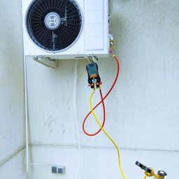 GOWENT SPÓŁKA Z OGRANICZONĄ ODPOWIEDZIALNOŚCIĄ - Biała jednostka zewnętrzna klimatyzatora Bosch zamontowana na ścianie, podłączona do manometru cyfrowego i butli z czynnikiem chłodniczym za pomocą czerwonych i żółtych przewodów. Widoczne...