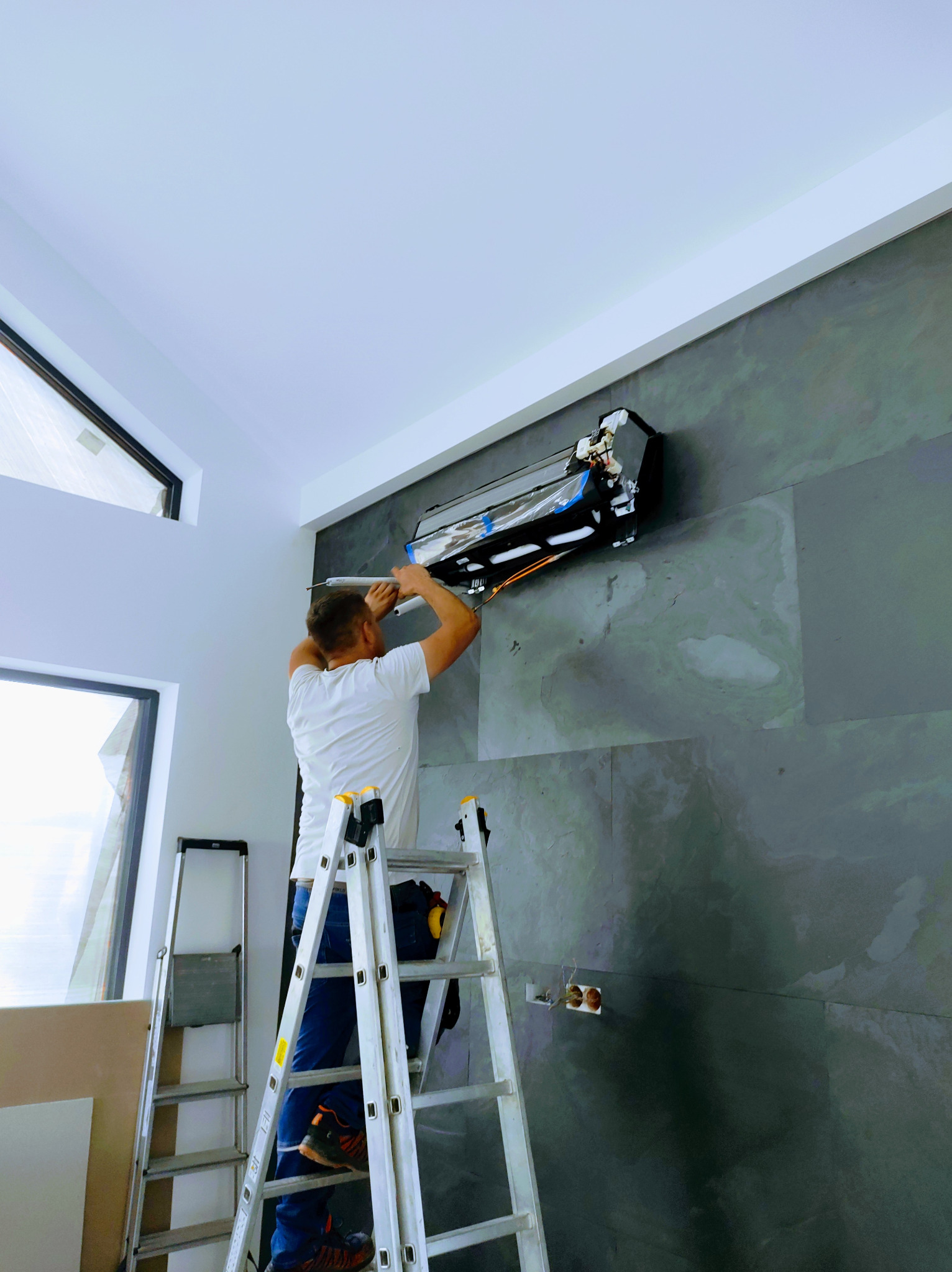 Instalator na drabinie podłącza przewody do klimatyzatora ściennego w nowoczesnym wnętrzu z oknami i szarą, kamienną ścianą.