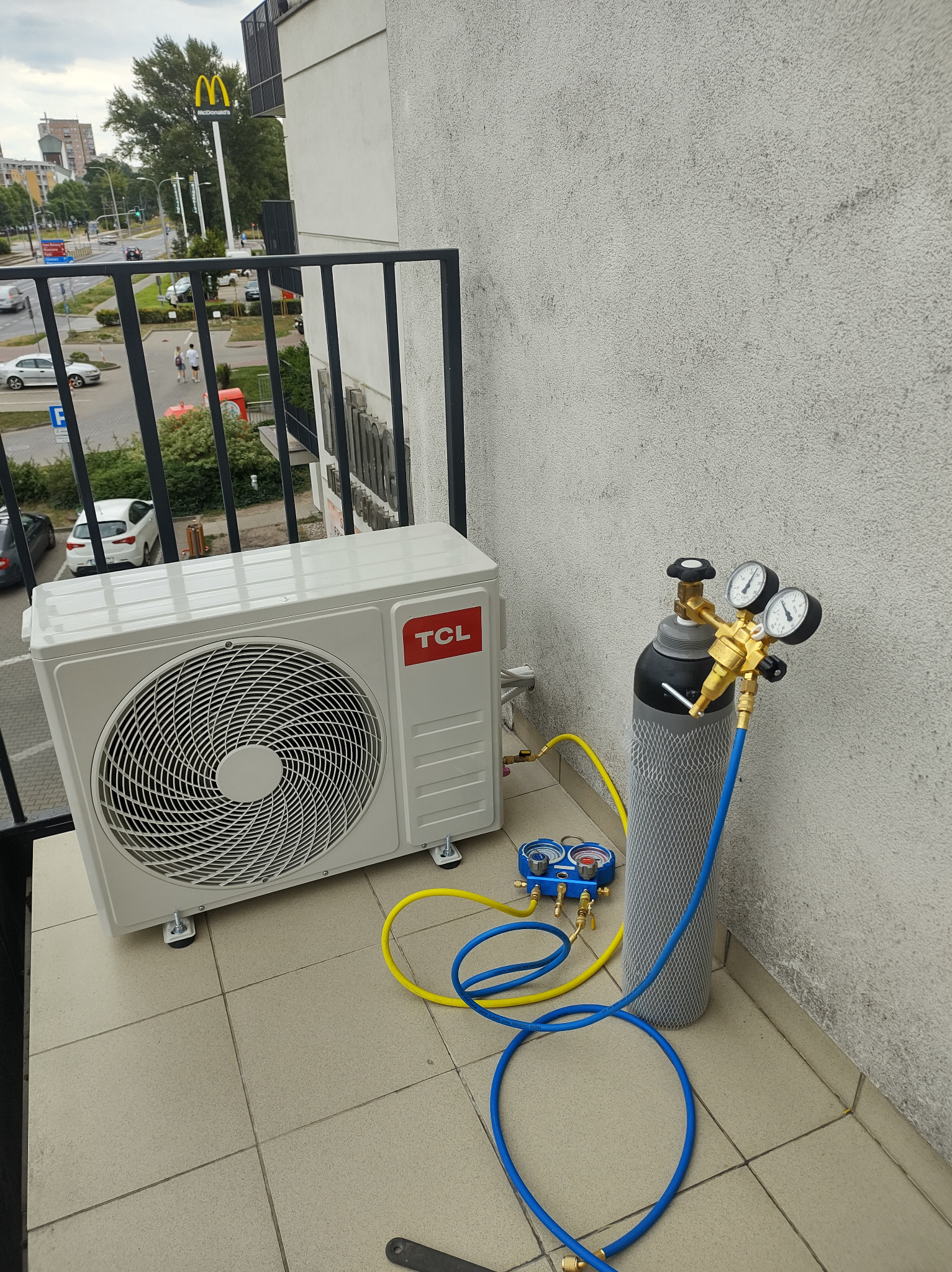 Biała jednostka zewnętrzna klimatyzacji TCL na balkonie, podłączona do butli z czynnikiem chłodniczym za pomocą żółtych i niebieskich węży oraz manometru.