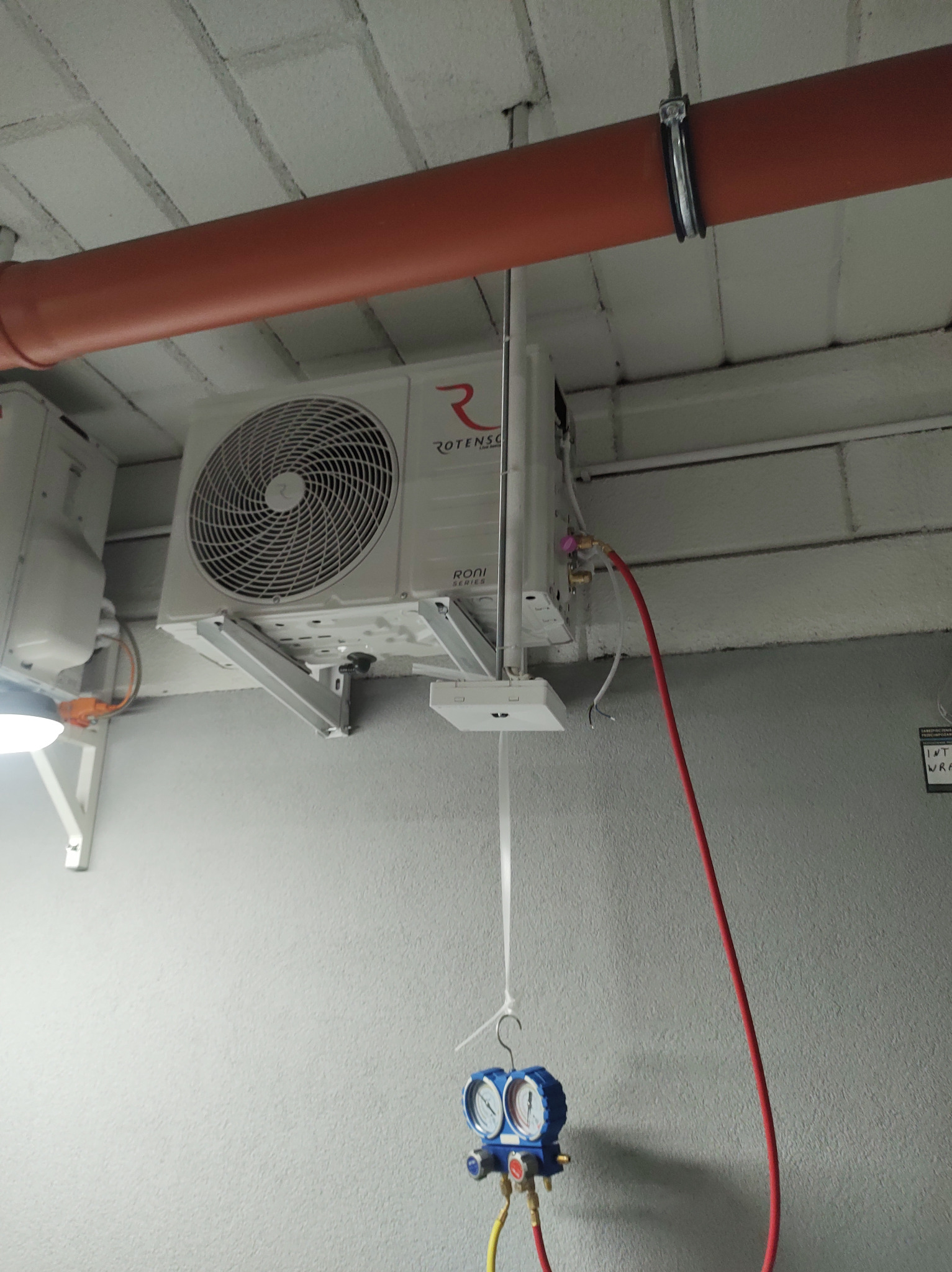 Biała jednostka wewnętrzna klimatyzacji marki Rotenso zamontowana pod sufitem, pod pomarańczową rurą. Podłączone są do niej przewody pomiarowe manometru w kolorach czerwonym i żółtym.