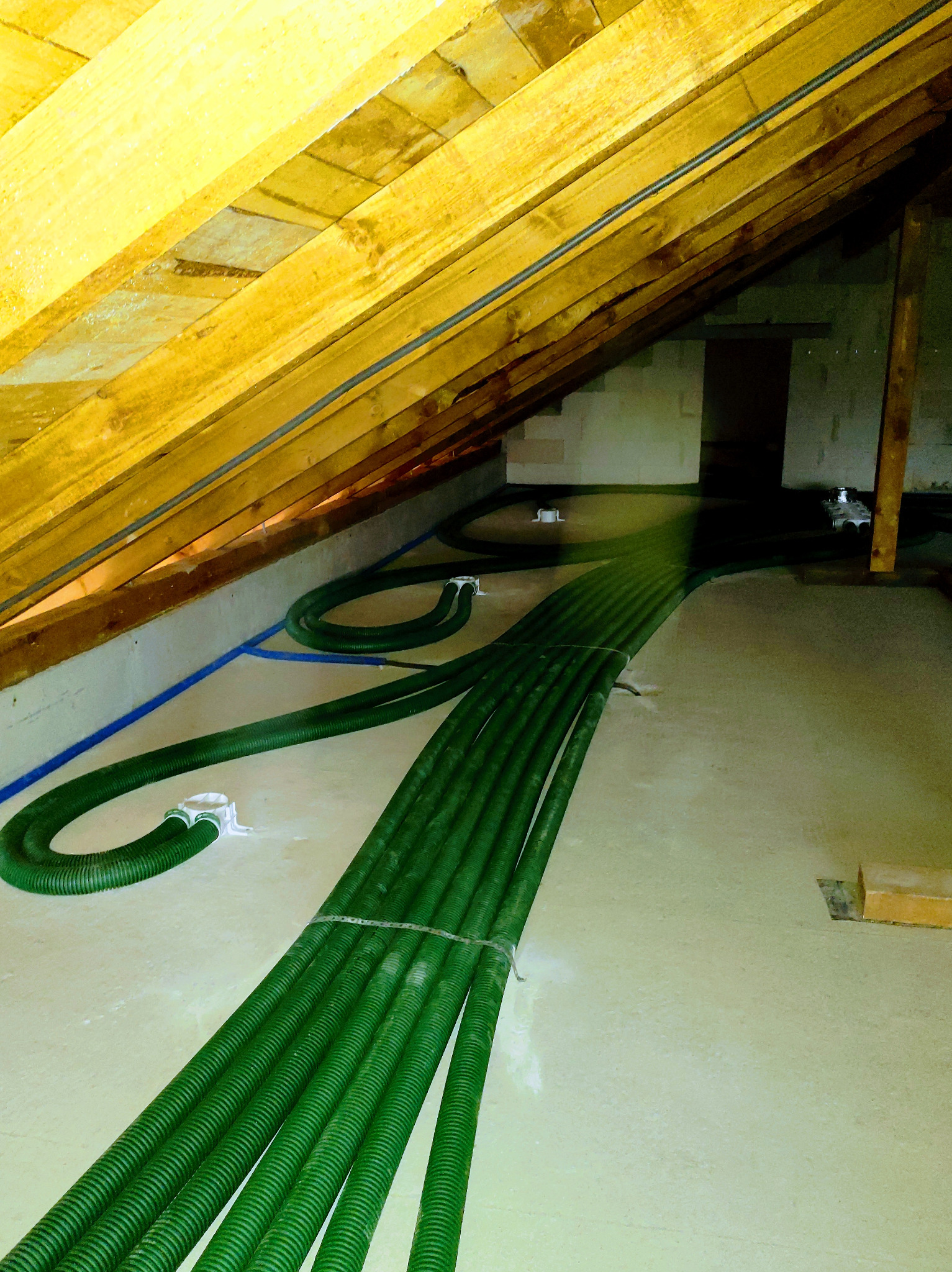 Zielone, karbowane rury wentylacyjne prowadzone w przestrzeni poddasza z drewnianą konstrukcją dachu i surową podłogą.