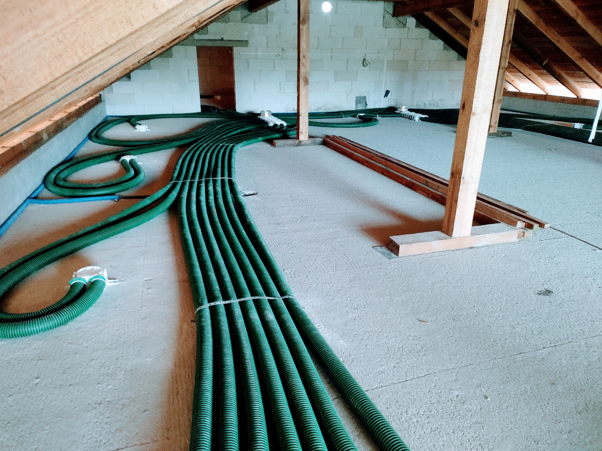 Poddasze w trakcie instalacji rekuperacji: widoczne zielone rury wentylacyjne, drewniane elementy konstrukcyjne dachu i ściany z betonu komórkowego.