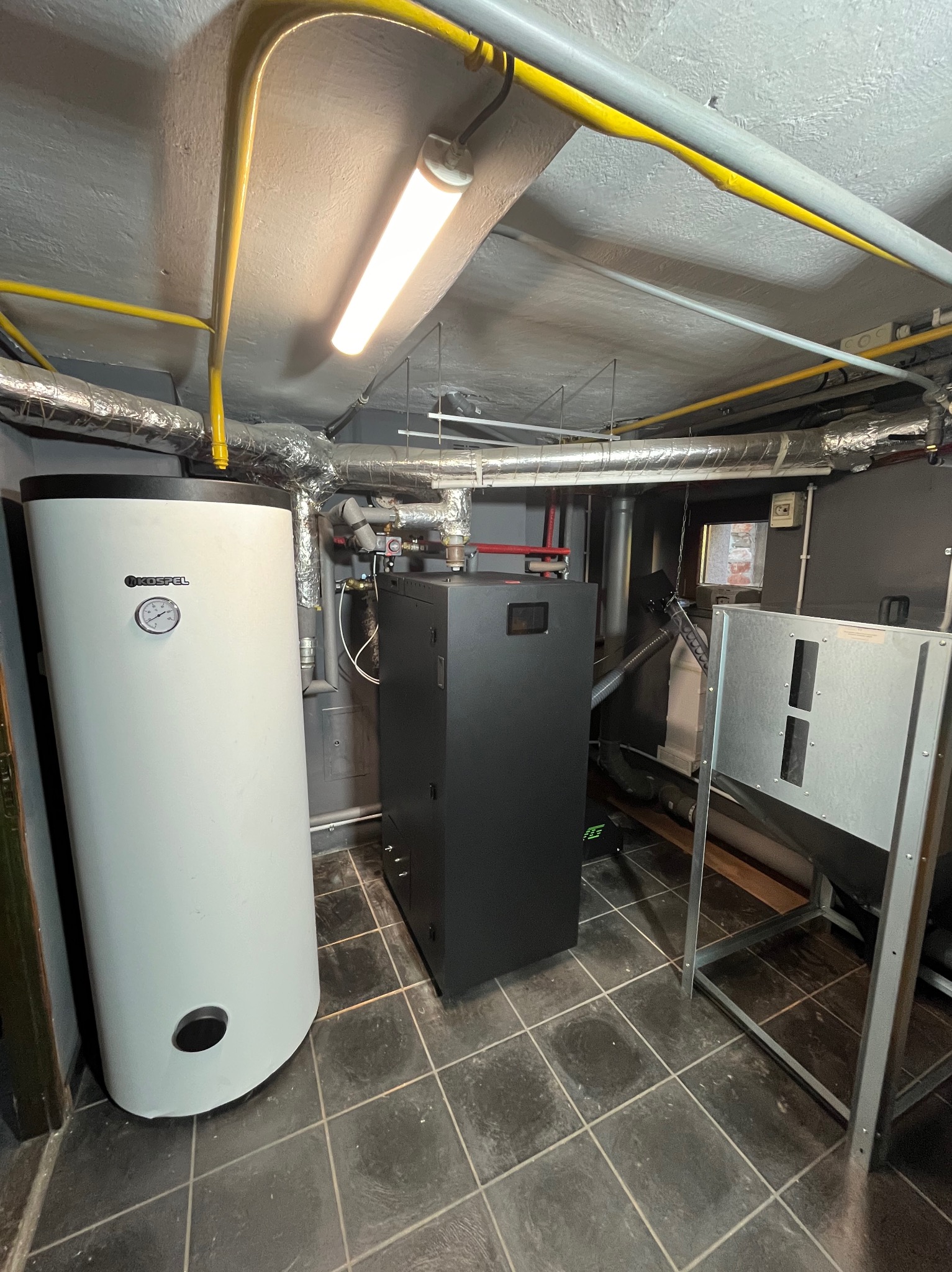 W kotłowni stoi biały zasobnik ciepłej wody Kospel oraz czarny piec. Widoczne rury instalacji grzewczej pod sufitem i fragment konstrukcji metalowej.