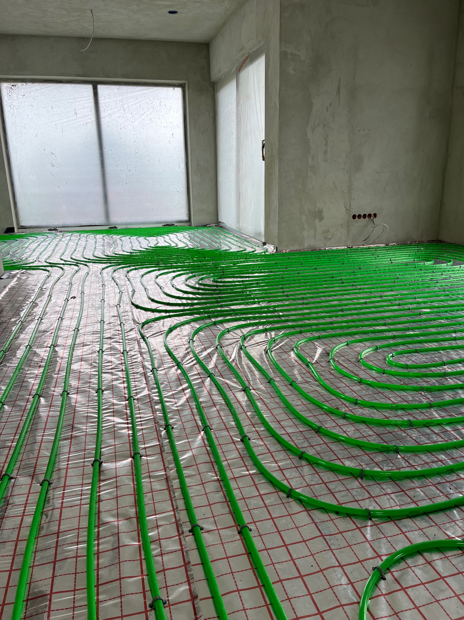 Instalacja ogrzewania podłogowego: zielone rury ułożone spiralnie na folii z czerwoną kratką w surowym wnętrzu z oknem