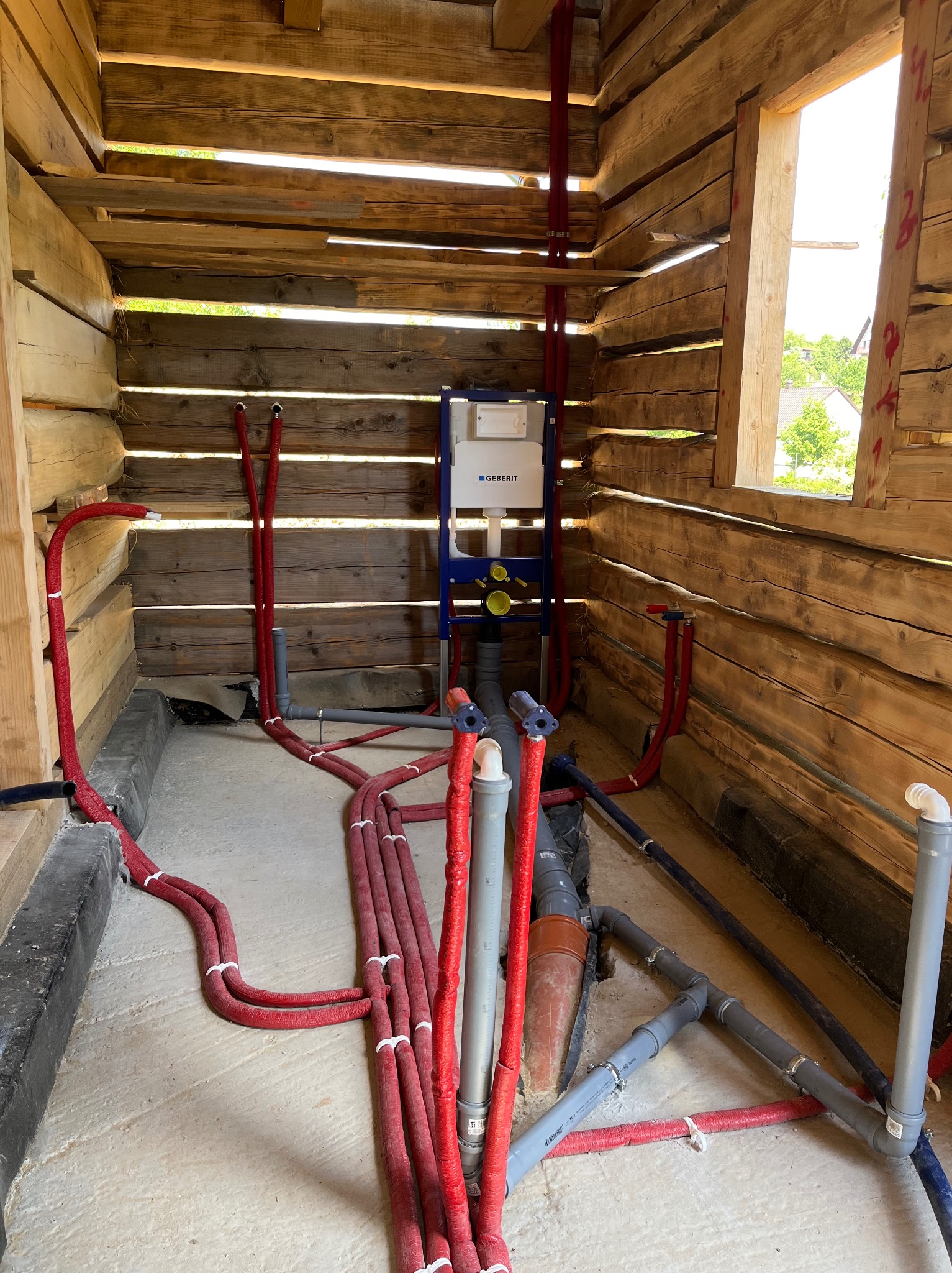 Surowe wnętrze drewnianego domu w trakcie instalacji hydraulicznej: widoczne rury w czerwonej izolacji, szare rury kanalizacyjne i stelaż podtynkowy WC marki Geberit. Przez okno widać zieleń.