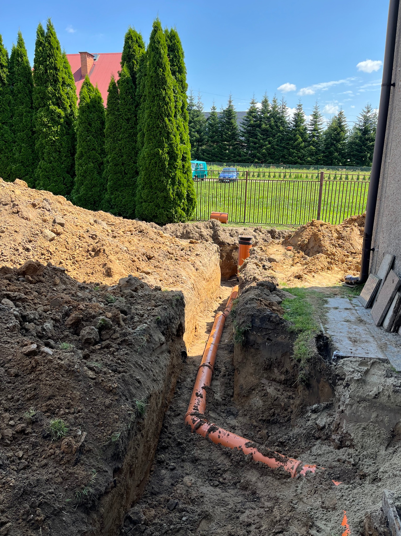 Wykop z ułożonym pomarańczowym rurociągiem kanalizacyjnym, widoczne połączenia rur, ziemia, trawa, w tle tuje i budynek.