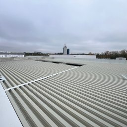 WillStall Patryk Wilczyński - Widok z góry na blaszany dach przemysłowy w trakcie montażu, z widocznymi elementami konstrukcyjnymi i pracownikiem w tle, na tle pochmurnego nieba i zabudowań miejskich.