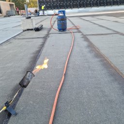 WillStall Patryk Wilczyński - Prace dekarskie: palnik dekarski w użyciu, widoczny płomień, butla gazowa i zgrzewane łączenie papy na dachu płaskim, w tle ekipa pracowników.