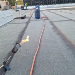 Prace dekarskie: palnik dekarski w użyciu, widoczny płomień, butla gazowa i zgrzewane łączenie papy na dachu płaskim, w tle ekipa pracowników.