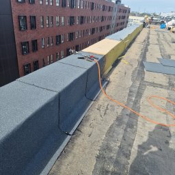 WillStall Patryk Wilczyński - Fragment dachu budynku z widoczną papą termozgrzewalną, narzędziami dekarskimi i pomarańczowym wężem, w tle budynek z czerwonej cegły i pracownicy na dachu