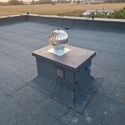 Płaski dach pokryty papą z zamontowanym kominkiem wentylacyjnym typu turbowent, widok na pola uprawne i dom jednorodzinny w oddali.