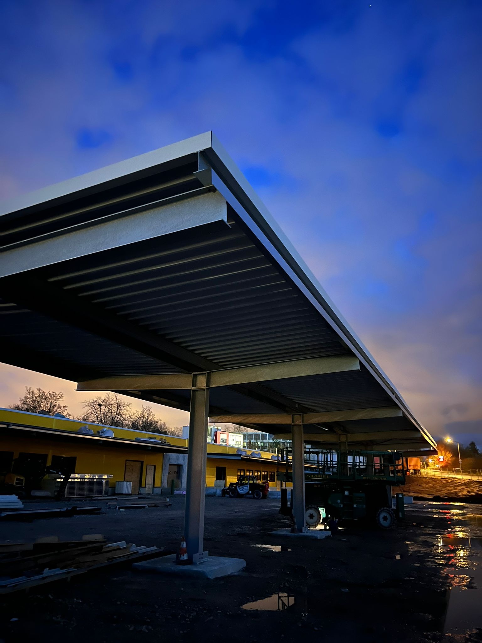 Konstrukcja zadaszenia z blachy trapezowej wsparta na metalowych słupach, widoczna na tle wieczornego, pochmurnego nieba i budynków w trakcie budowy.