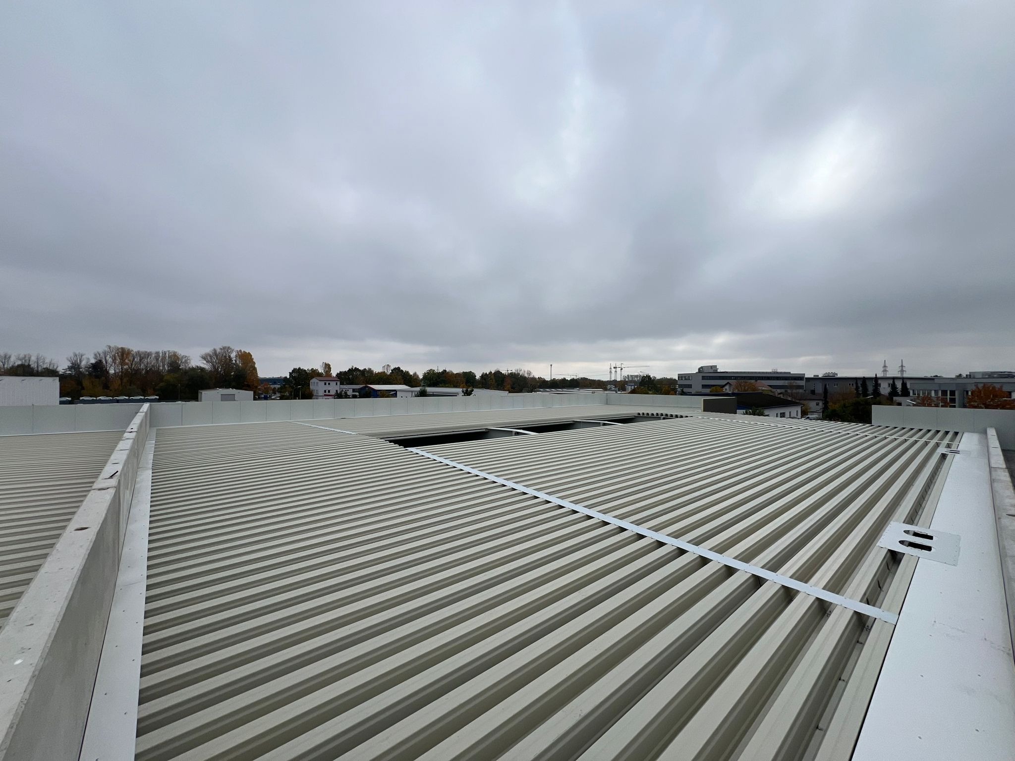 Widok z góry na płaski dach przemysłowy z blachy trapezowej, częściowo zamontowany, z widocznymi elementami mocującymi i fragmentem niezadaszonym, na tle pochmurnego nieba i zabudowań.