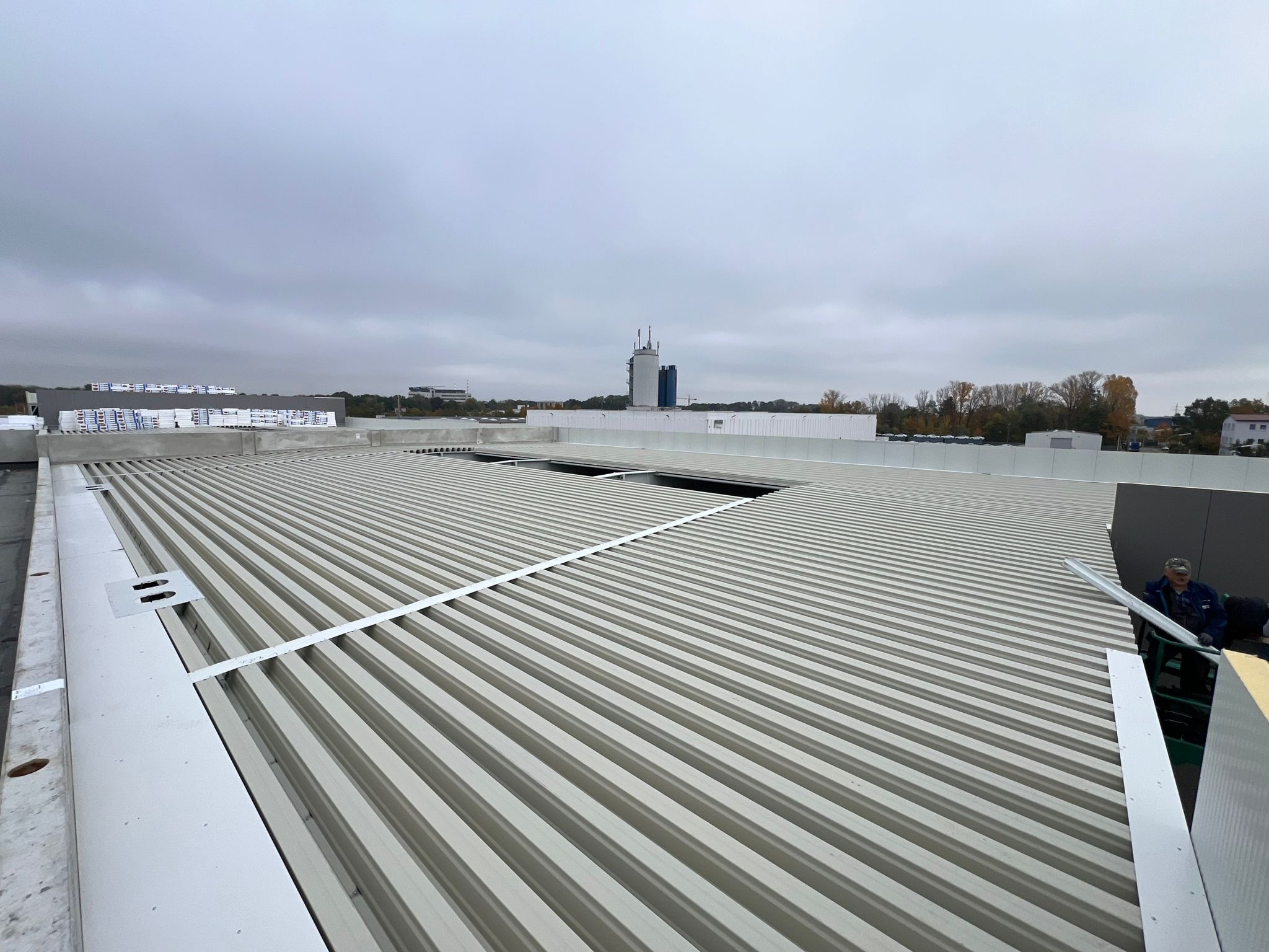 Widok z góry na blaszany dach przemysłowy w trakcie montażu, z widocznymi elementami konstrukcyjnymi i pracownikiem w tle, na tle pochmurnego nieba i zabudowań miejskich.