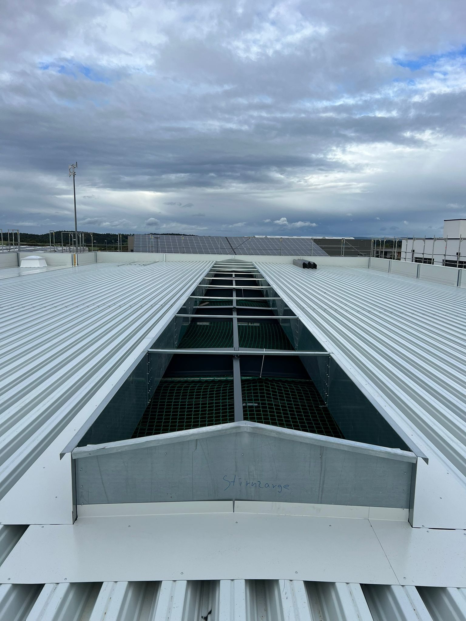 Widok z góry na płaski dach z blachy trapezowej z otworem, zabezpieczonym siatką, w tle panele słoneczne i pochmurne niebo.