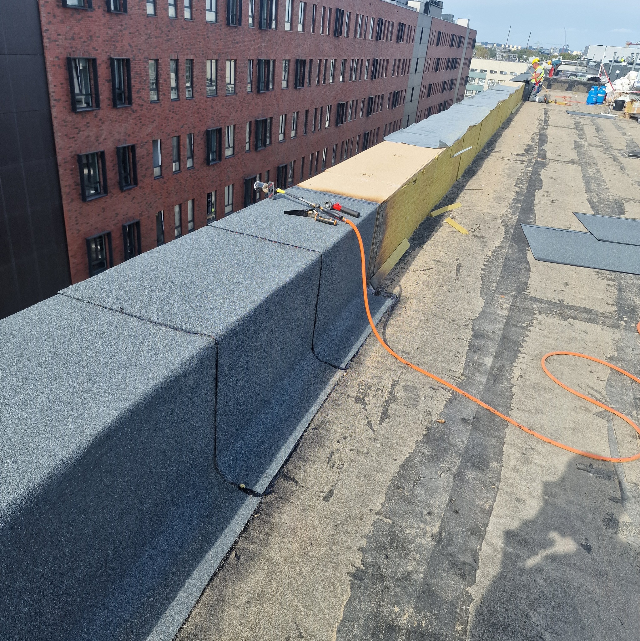 Fragment dachu budynku z widoczną papą termozgrzewalną, narzędziami dekarskimi i pomarańczowym wężem, w tle budynek z czerwonej cegły i pracownicy na dachu