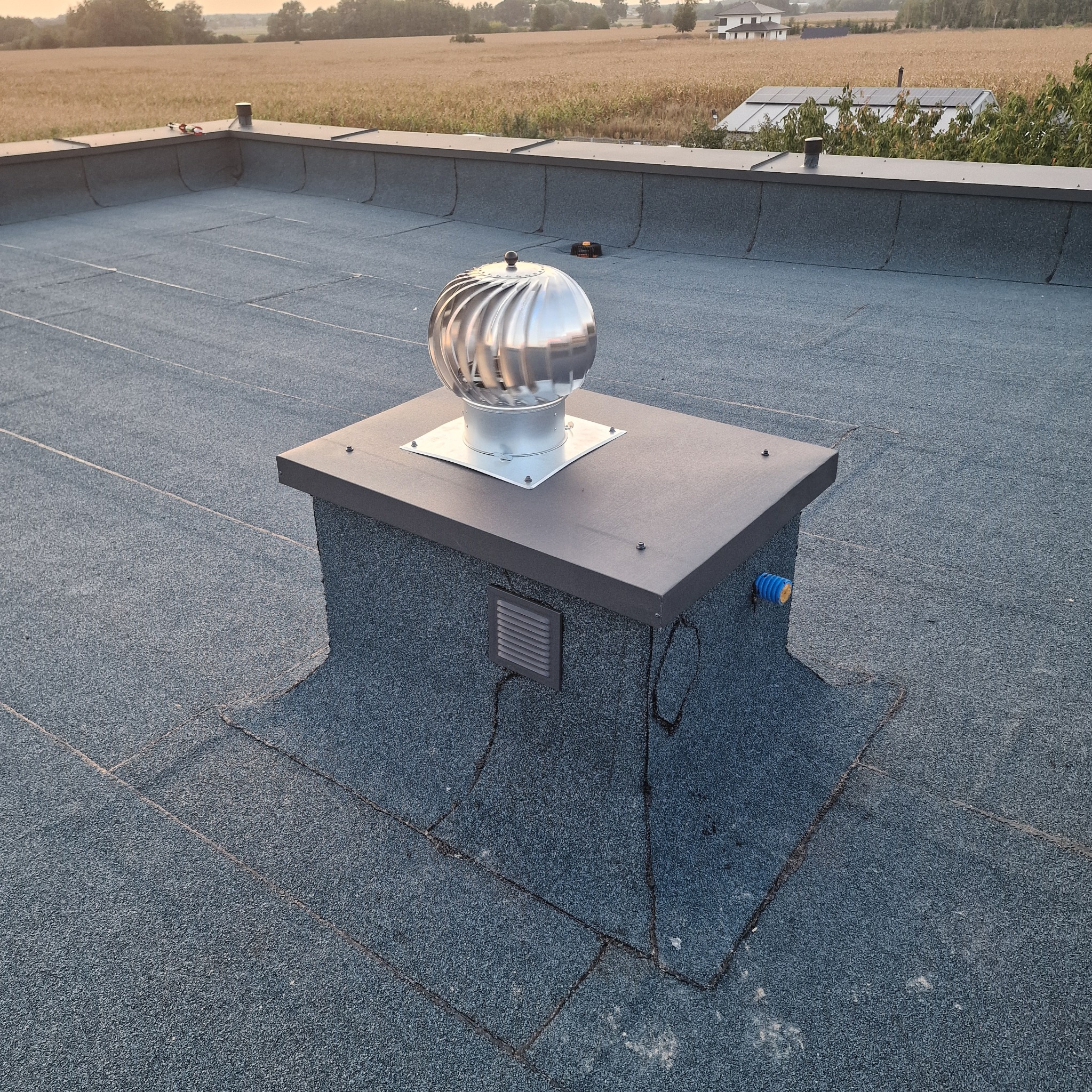 Płaski dach pokryty papą z zamontowanym kominkiem wentylacyjnym typu turbowent, widok na pola uprawne i dom jednorodzinny w oddali.