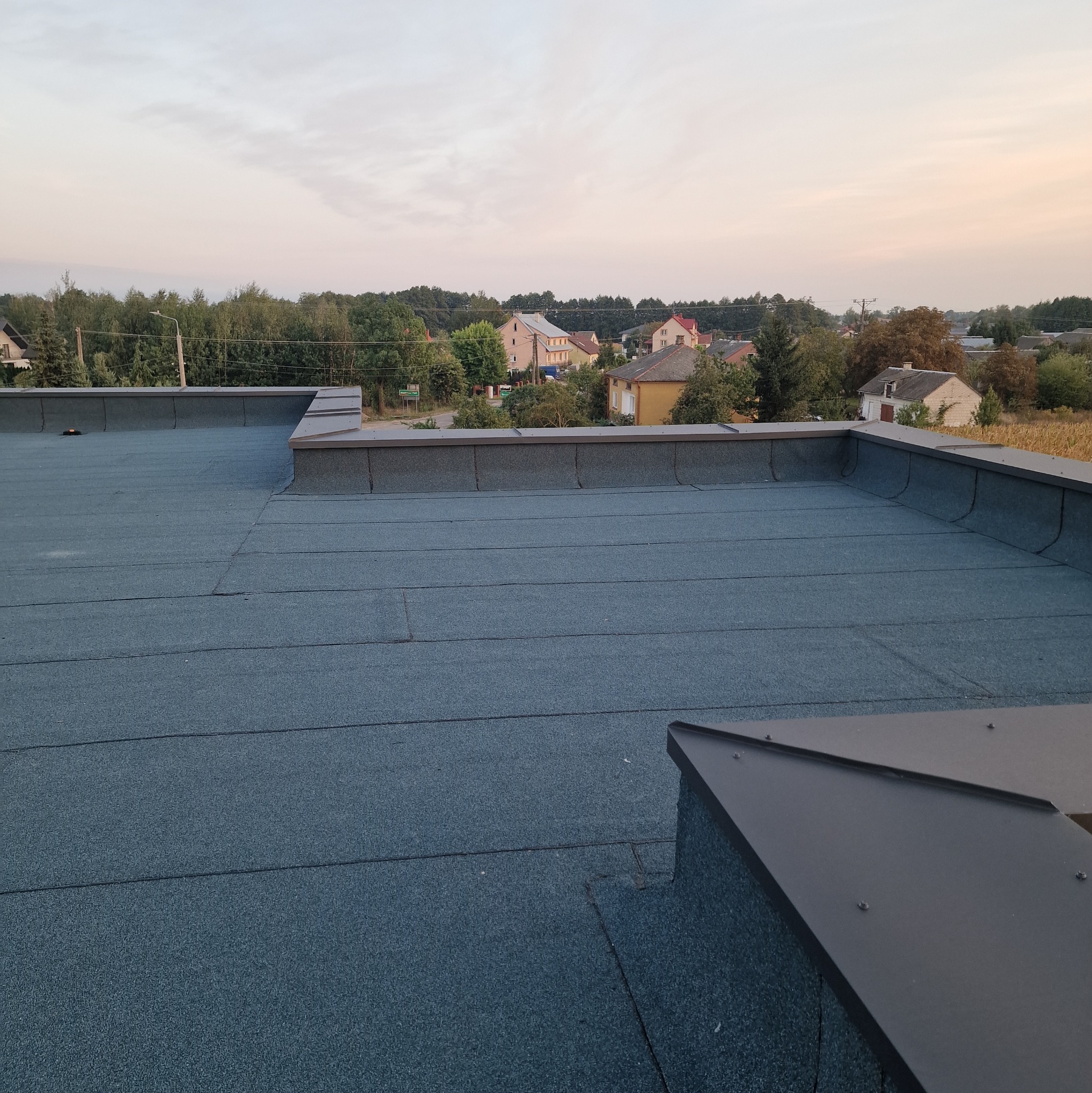 Widok z góry na płaski dach pokryty papą termozgrzewalną, z widocznymi obróbkami blacharskimi i zabudową attyki na tle zabudowań wiejskich i zachodzącego słońca.