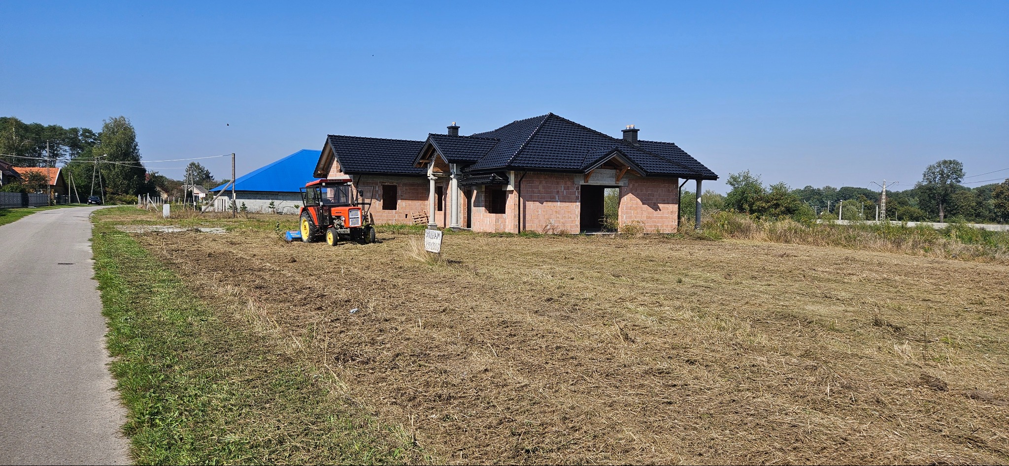 Traktor z kosiarką rotacyjną na polu po skoszeniu trawy, w tle niedokończony dom z cegły i błękitny dach budynku gospodarczego.