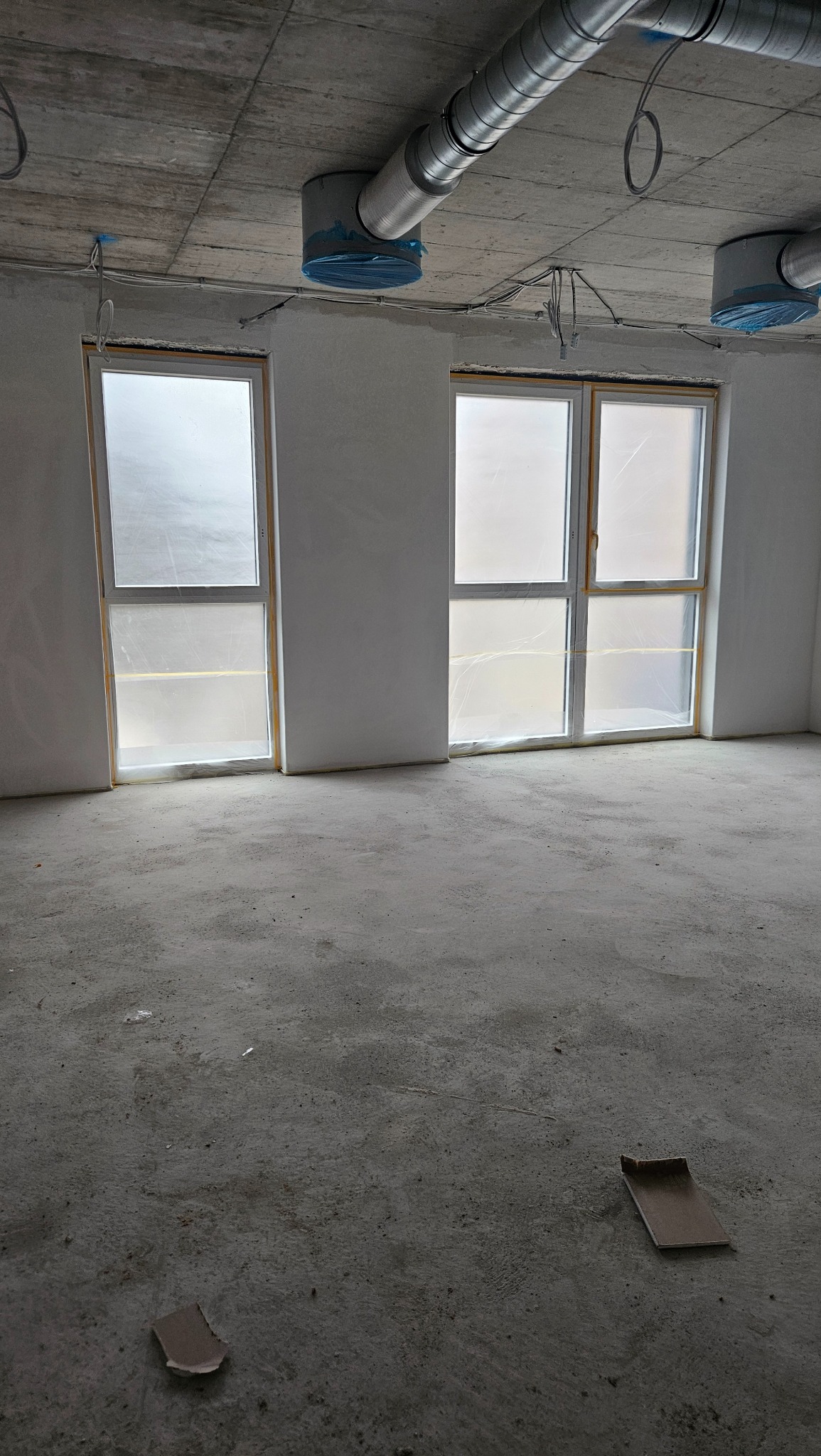 Surowe wnętrze w trakcie wykańczania: dwa okna z folią ochronną, betonowa podłoga, sufit z odsłoniętą instalacją wentylacyjną i przewodami elektrycznymi.