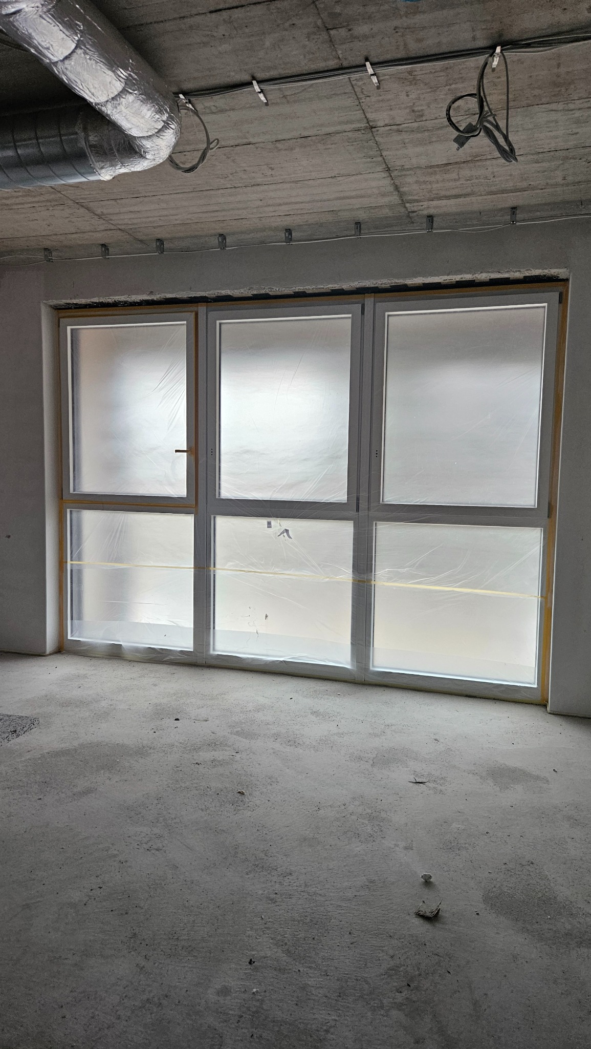 Trzyczęściowe białe okno z szybami zabezpieczonymi folią ochronną w surowym wnętrzu, widoczne przewody i rury wentylacyjne pod betonowym sufitem.