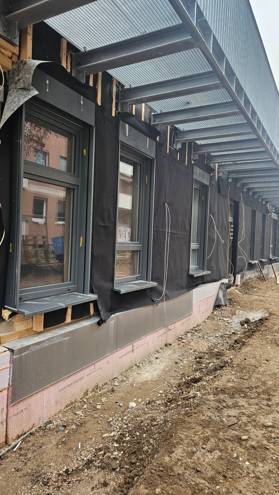 Montaż okien w nowym budynku z widocznym ociepleniem fundamentów i metalową konstrukcją dachu nad oknami. Czarna membrana izolacyjna wokół ram okiennych.