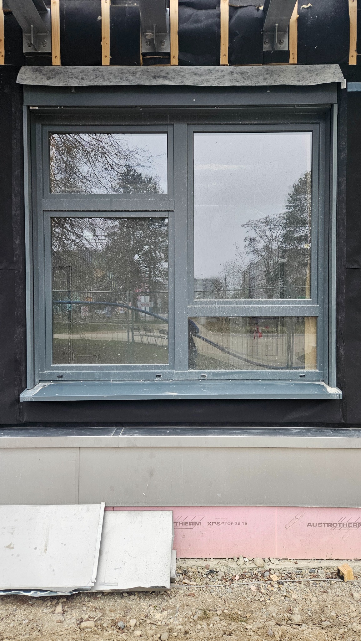 Szare okno z roletą zewnętrzną w trakcie montażu, widoczne elementy konstrukcyjne i izolacyjne budynku.