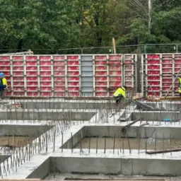 Wykopany i zalany betonem fundament z wystającymi prętami zbrojeniowymi, widoczne szalunki w kolorze czerwonym, pracownicy w odblaskowych kamizelkach i kaskach na budowie.